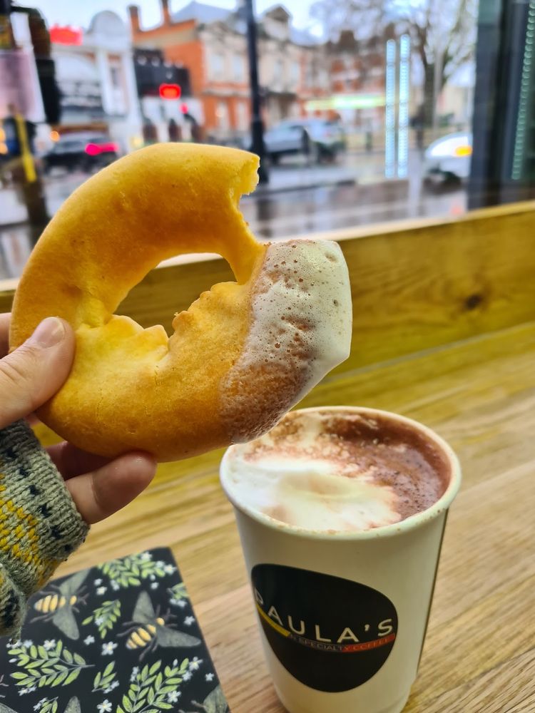 A ring of Colombian bread being dunked into a cup of frothy hot chocolate on the window bar, with Brixton Road outside