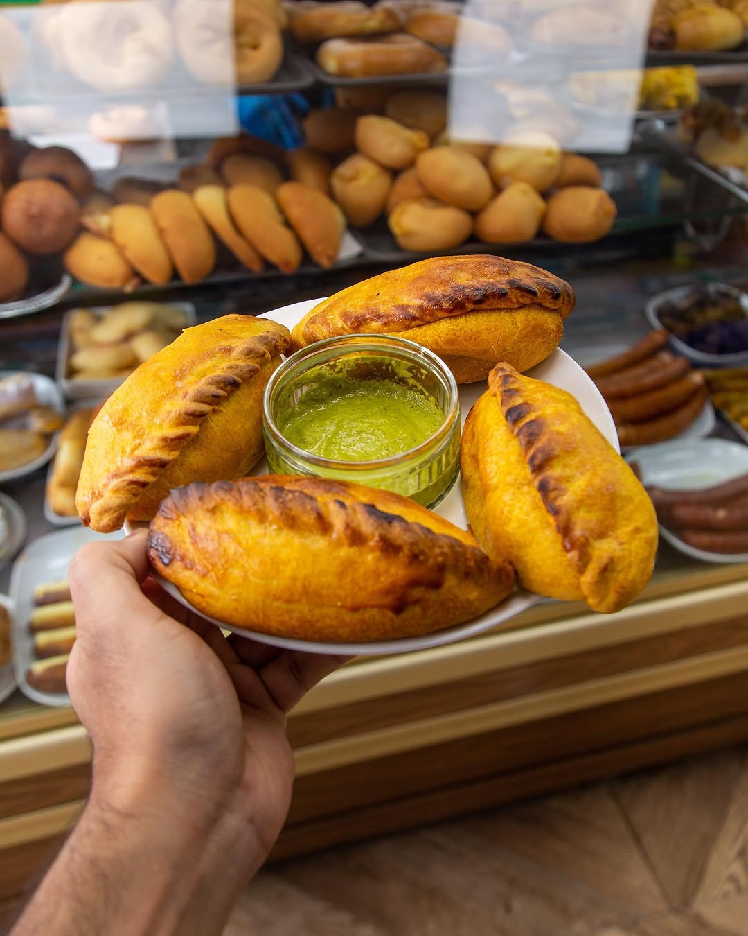 A plate of golden empanadas held up in front of the counter, with a small glass pot of green chilli sauce in the middle
