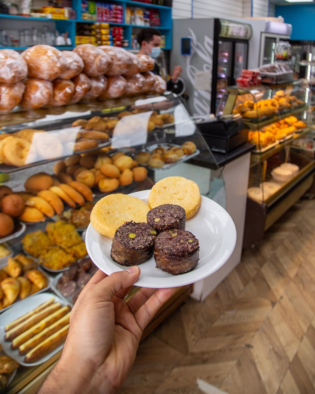 A plate of arepas and Colombian black pudding held up in front of the shop's cabinets of breads and pastries