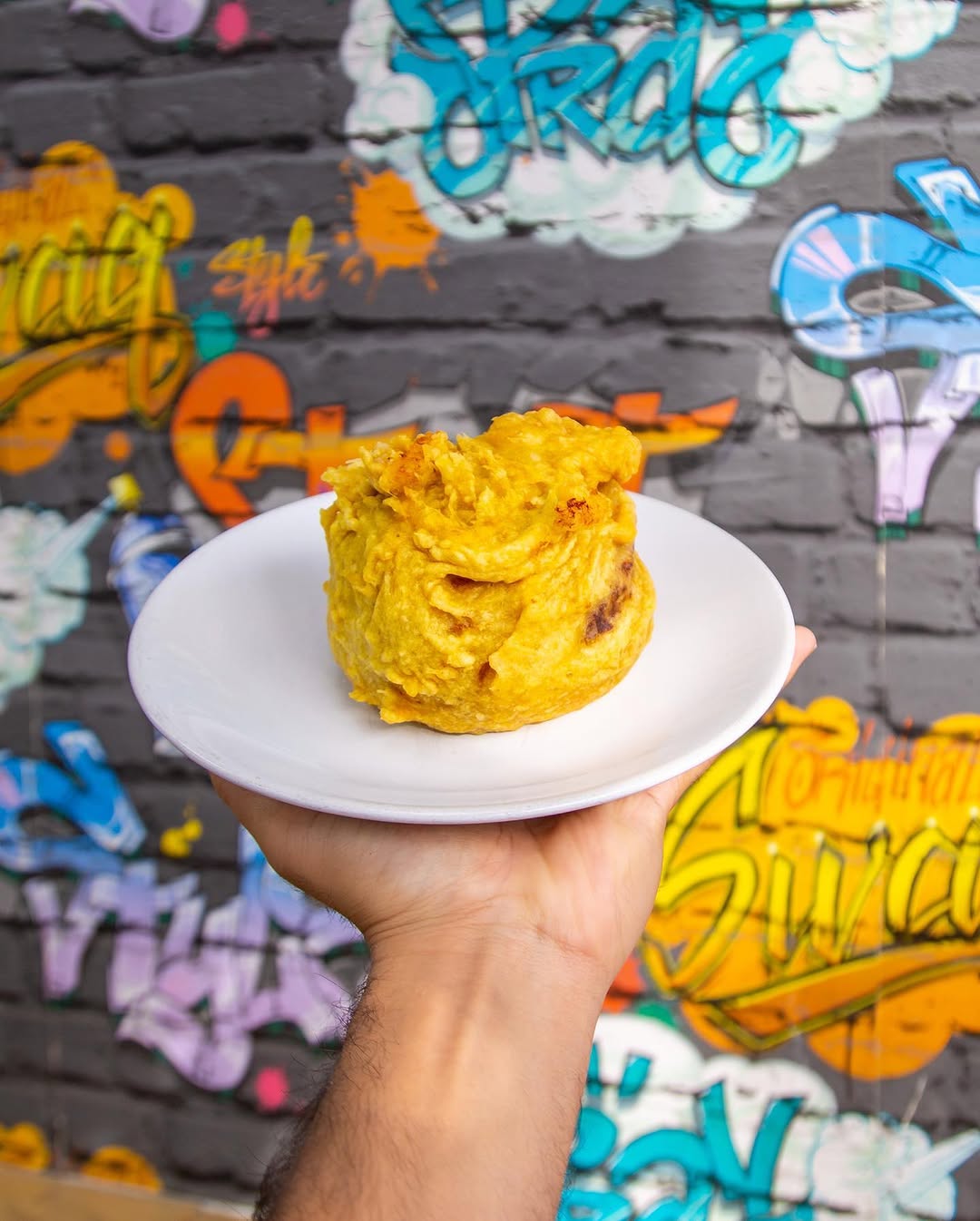 A papa rellena on a white plate, held up against the painted brick wall outside the shop